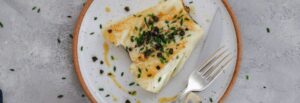 An up close image of pan seared seabass with capers on a white plate with a silver fork.