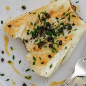 An up close image of pan seared seabass with capers on a white plate with a silver fork.