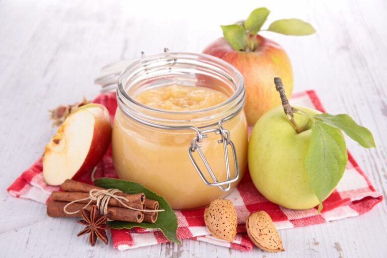 An up close image of a mason jar filled with applesauce, surrounded by apples, pears, and cinnamon sticks on a checkered red and white cloth