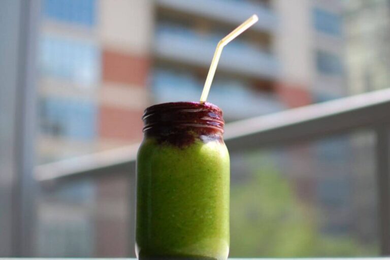 A close up image of a green smoothie with a straw that is in front of a blurred city landscape