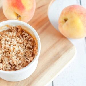 A close up image of a white ramekin that is filled with the peach crisp, on a cutting board sitting atop of a white wooden table that has two fresh peaches on top.