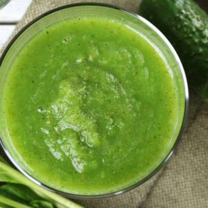 An up close image of a green smoothie in a glass next to a cucumber and spinach