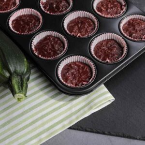 A dark countertop displays a muffin tray filled with glossy, deep-red mixture in each cup. Adjacent to the tray is a fresh, green zucchini with a striped cloth beneath. A small sprig of herbs lies at the upper right corner of the image.