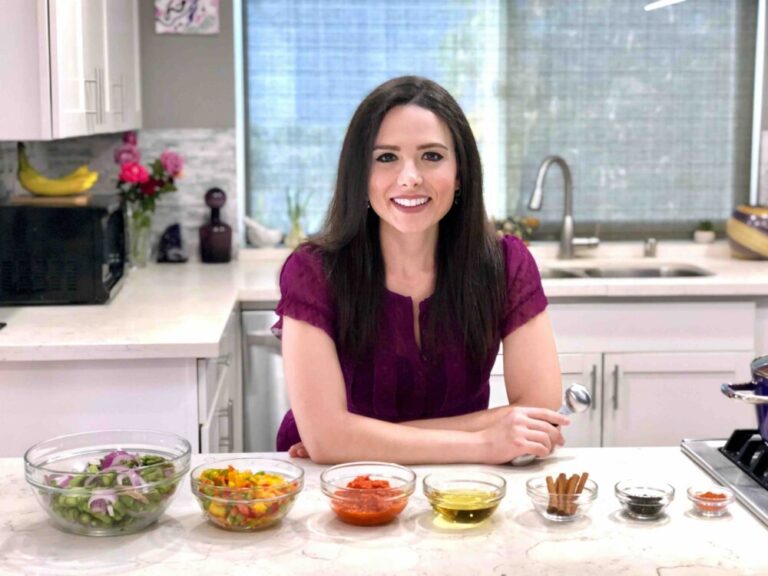 Smiling woman sitting at a kitchen counter with various ingredients in clear bowls, ready for cooking. She's holding a measuring spoon and appears to be giving a cooking demonstration. The kitchen is modern with a white countertop, and there are pink flowers and a fruit bowl in the background.