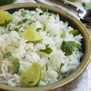 A bowl of cilantro lime basmati rice garnished with lime wedges and fresh cilantro leaves on a rustic wooden table. The rice is fluffy and white, and the bright green of the cilantro adds a fresh contrast. The lime wedges are arranged on top of the rice. A spoon and a folded napkin are placed beside the bowl, indicating readiness for serving. The background is softly focused, emphasizing the dish.