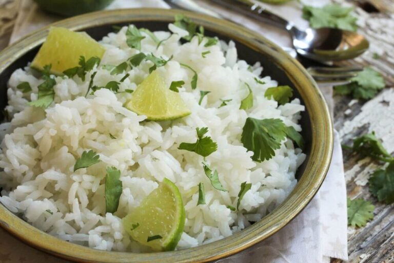 A bowl of cilantro lime basmati rice garnished with lime wedges and fresh cilantro leaves on a rustic wooden table. The rice is fluffy and white, and the bright green of the cilantro adds a fresh contrast. The lime wedges are arranged on top of the rice. A spoon and a folded napkin are placed beside the bowl, indicating readiness for serving. The background is softly focused, emphasizing the dish.