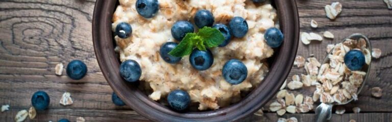 A bowl of oatmeal topped with blueberries and a sprig of mint on a wooden table. Beside the bowl is a silver spoon with oat flakes and a blueberry on top. The table has scattered oat flakes and blueberries around the bowl.