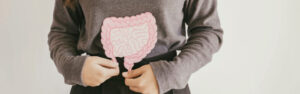 This image shows a person holding a cut-out illustration of a human intestine in front of their abdomen. The person is wearing a gray long-sleeved top and is standing against a neutral background. The intestine illustration is detailed, showing the complex looping and folding typical of the intestinal tract, and is colored in shades of pink and white, possibly to indicate the various layers and textures of the organ. The person's face is not visible in the frame, focusing the viewer's attention on the intestine model they are holding.