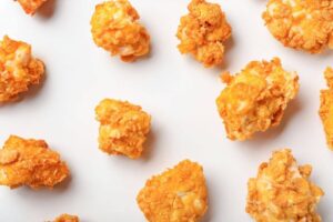 An array of golden-brown chicken nuggets scattered across a white background. Each nugget has a crispy coating with a visibly textured surface, suggesting a crunchy bite. The golden color varies slightly from piece to piece, with some lighter and darker spots, indicating they have been baked to a delectable crispness.