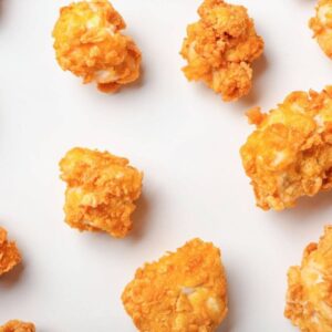 An array of golden-brown chicken nuggets scattered across a white background. Each nugget has a crispy coating with a visibly textured surface, suggesting a crunchy bite. The golden color varies slightly from piece to piece, with some lighter and darker spots, indicating they have been baked to a delectable crispness.