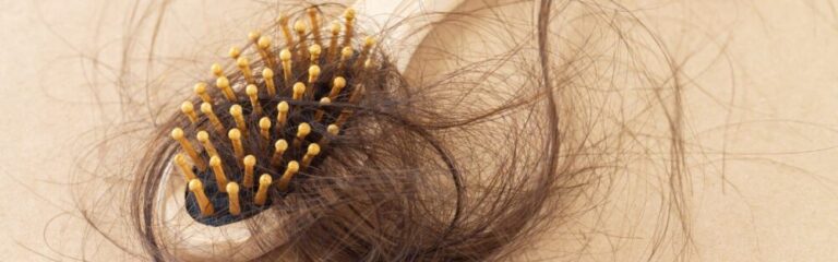 An image showing a close-up view of a hairbrush entangled with strands of brown hair. The brush has a navy-blue base with an array of rounded, wooden tips on bristles. Some hair is wrapped around the bristles, while loose strands drape over the edge of the brush, against a beige background.