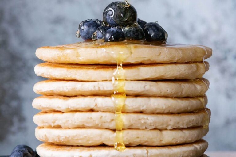 Stack of golden-brown oat pancakes topped with fresh blueberries and drizzled with golden maple syrup on a light-textured background.