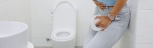 A person experiencing discomfort stands next to a toilet, clutching their abdomen with one hand and holding a roll of toilet paper in the other, suggesting the urgency and discomfort associated with conditions like ulcerative colitis.