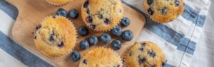 A wooden board topped with freshly baked gluten-free lemon blueberry muffins. The muffins are golden brown with visible blueberries scattered throughout. They are surrounded by a few loose blueberries, and the board is placed on a blue and white striped cloth. These muffins are a delicious and suitable option for those with inflammatory bowel disease (IBD).