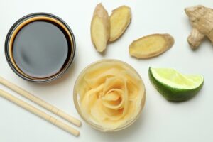 Ingredients for a 3-ingredient ginger lime dressing displayed on a light surface: a small bowl of coconut aminos, several slices of fresh ginger, a lime wedge, and a glass bowl filled with thinly sliced ginger. Wooden chopsticks rest next to the coconut aminos bowl.