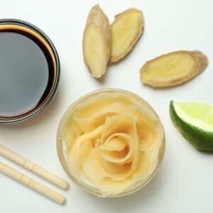 Ingredients for a 3-ingredient ginger lime dressing displayed on a light surface: a small bowl of coconut aminos, several slices of fresh ginger, a lime wedge, and a glass bowl filled with thinly sliced ginger. Wooden chopsticks rest next to the coconut aminos bowl.