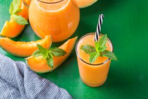 A refreshing glass of cantaloupe agua fresca garnished with a sprig of mint and served with a striped straw. The drink is a vibrant orange color, poured into a glass with a pitcher of more cantaloupe agua fresca in the background. Fresh cantaloupe slices are also visible around the pitcher and glass, placed on a green surface next to a textured gray napkin.