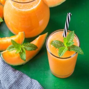 A refreshing glass of cantaloupe agua fresca garnished with a sprig of mint and served with a striped straw. The drink is a vibrant orange color, poured into a glass with a pitcher of more cantaloupe agua fresca in the background. Fresh cantaloupe slices are also visible around the pitcher and glass, placed on a green surface next to a textured gray napkin.