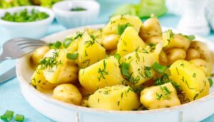 A bowl of healthy herbed potato salad featuring chunks of boiled potatoes garnished with fresh dill and chopped green onions. The salad is served in a white dish with a fork resting on the side. Small bowls of additional green onions and herbs are visible in the background, along with a light blue tablecloth and fresh green vegetables, creating a vibrant and appetizing presentation.