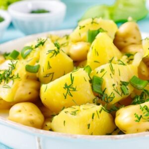 A bowl of healthy herbed potato salad featuring chunks of boiled potatoes garnished with fresh dill and chopped green onions. The salad is served in a white dish with a fork resting on the side. Small bowls of additional green onions and herbs are visible in the background, along with a light blue tablecloth and fresh green vegetables, creating a vibrant and appetizing presentation.
