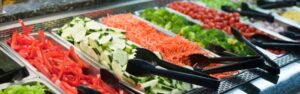 A close-up view of a fresh salad bar featuring a colorful array of sliced vegetables, including red bell peppers, cucumbers, shredded carrots, broccoli, and cherry tomatoes, neatly arranged in metal containers with black serving tongs. This image highlights the variety and freshness typically found in salads, which may be a consideration for those with Crohn's disease.