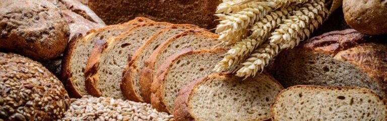 An assortment of rustic whole-grain breads, including sliced and whole loaves, arranged with golden wheat stalks and scattered grains. The bread has a crispy, golden-brown crust and a soft, airy interior, highlighting its artisanal quality.