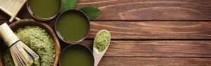 Top view of traditional matcha tea setup on a rustic wooden surface, featuring bowls of prepared matcha tea, a wooden spoon and bowl filled with green matcha powder, a bamboo whisk (chasen), and green leaves for decoration.
