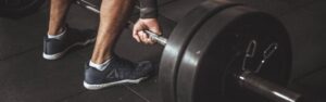 Close-up of a person gripping a loaded barbell, preparing to deadlift in a gym. The individual wears athletic shoes and stands on a rubber gym floor, emphasizing strength and focus.