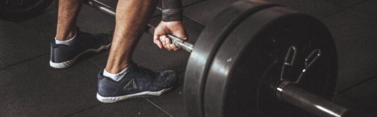 Close-up of a person gripping a loaded barbell, preparing to deadlift in a gym. The individual wears athletic shoes and stands on a rubber gym floor, emphasizing strength and focus.