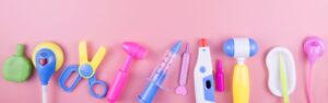 Brightly colored toy medical instruments, including a syringe, thermometer, scissors, and reflex hammer, lined up on a pink background representing pediatric healthcare and children’s medical care for Crohn's disease.